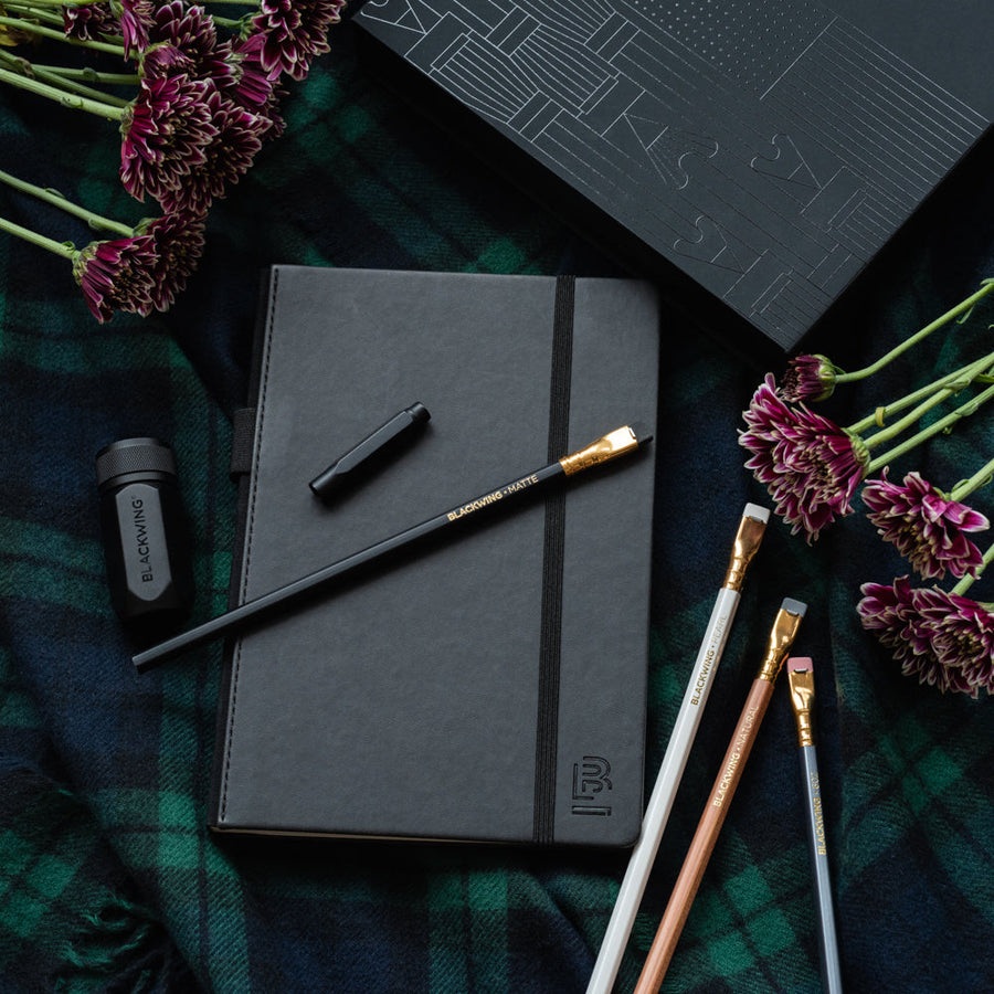 A Blackwing Notebook Essentials Set, Blackwing pencils, and flowers on a plaid blanket.