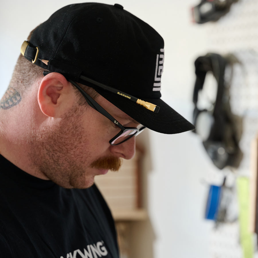 man wearing a black cap with a white embroidered Blackwing emblem on the front, featuring a pencil loop on the side holding a Blackwing Matte pencil