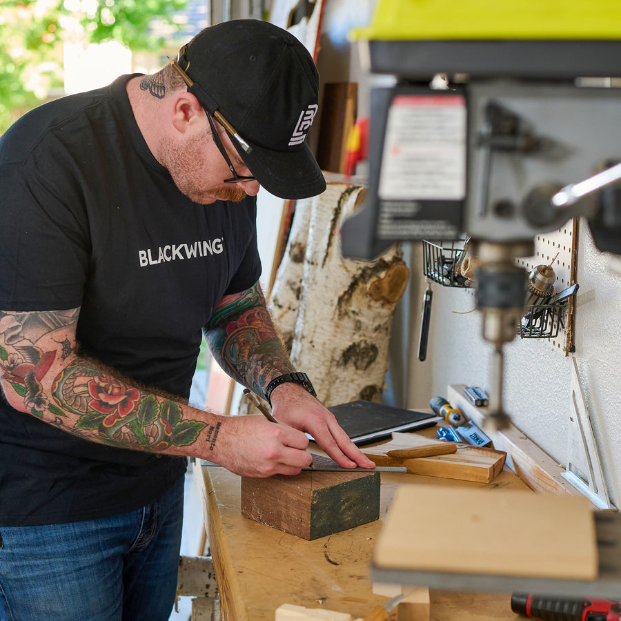 man doing woodworking in his shop, using a Blackwing Volume 21 pencil to make markings on wood. He is wearing a black canvas hap with a white Blackwing emblem embroidered on the front