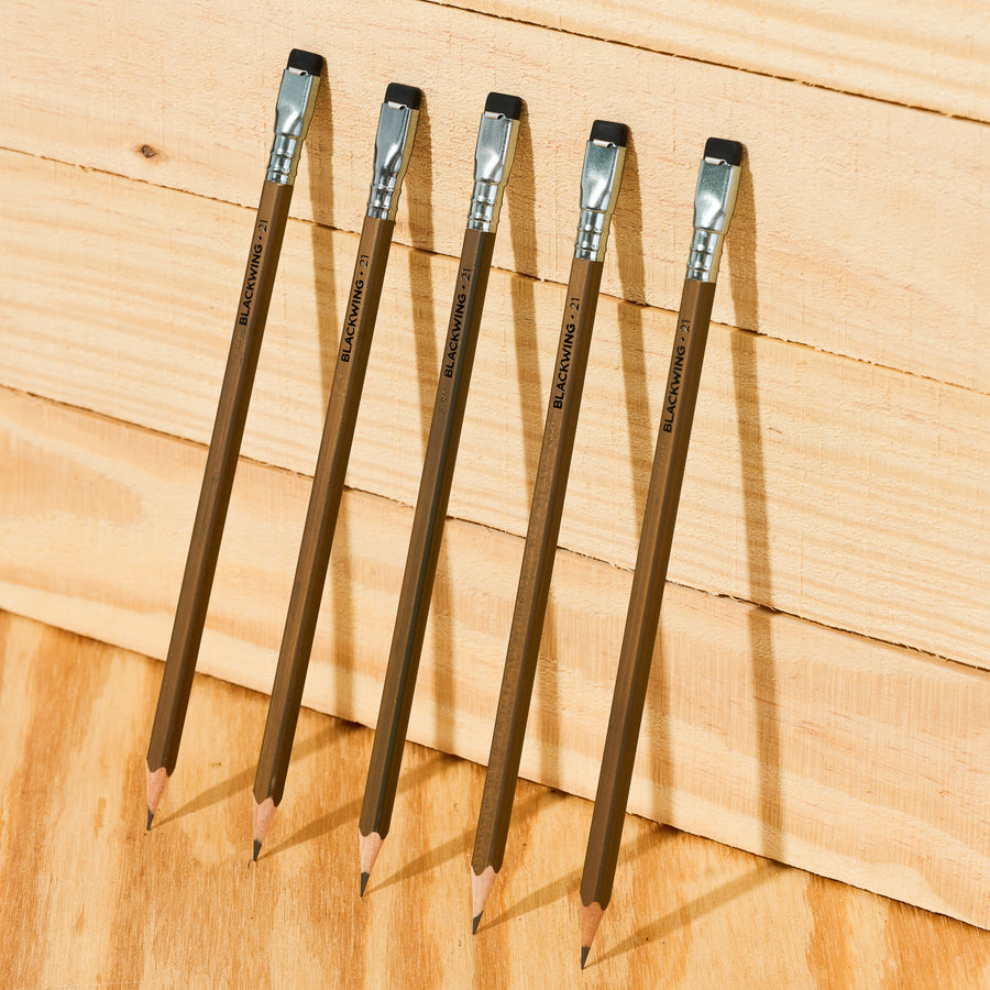 five Blackwing Volume 21 pencils leaning against a wood wall. These pencils feature a walnut-stain barrel, silver ferrule, black eraser, and black imprint